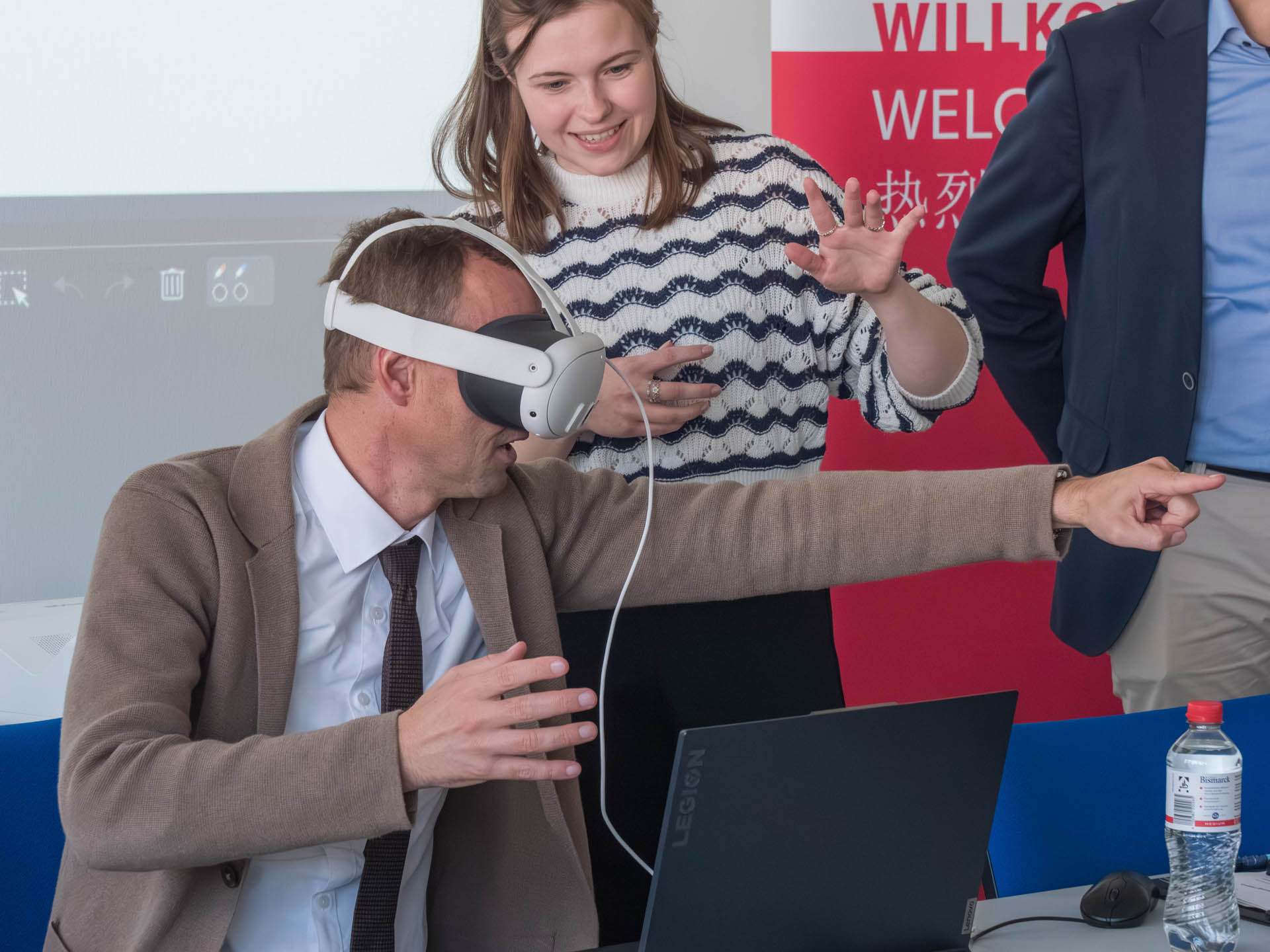 Ein Mann trägt eine VR-Brille und zeigt nach vorne, dahinter steht eine Frau, die etwas erklärt